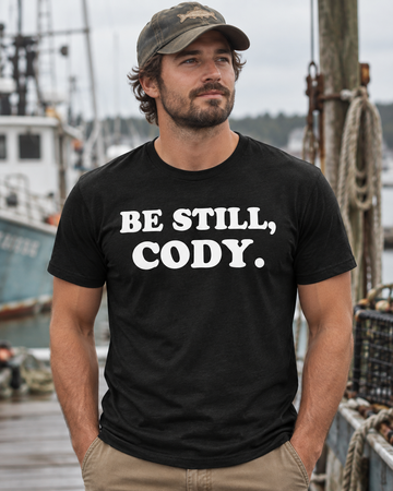 AI-generated lifestyle visualization: person wearing the black tri-blend Be Still, Cody. quote T-shirt in a sun-faded marina repair dock with ropes, buoys, and calm morning water
