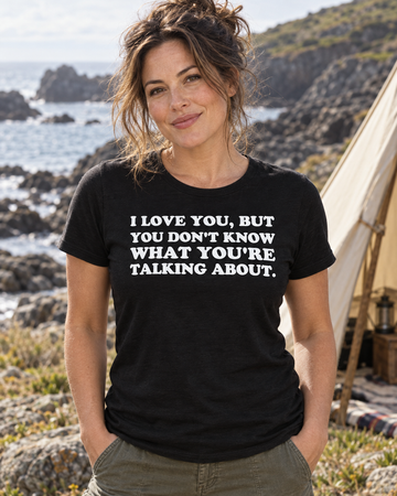 AI-generated lifestyle visualization: person wearing the black tri-blend I Love You, but You Don't Know What You're Talking About. quote T-shirt in a rocky coastal inlet with a small canvas tent, pale summer sky, and balanced storybook framing
