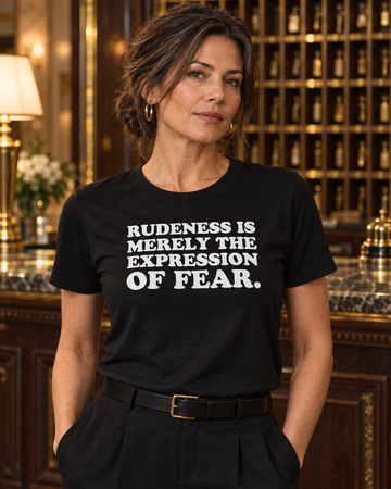AI-generated lifestyle visualization: person wearing the black tri-blend Rudeness Is Merely the Expression of Fear. quote T-shirt in an ornate hotel concierge desk with key cubbies, burgundy uniforms nearby, and warm chandelier light