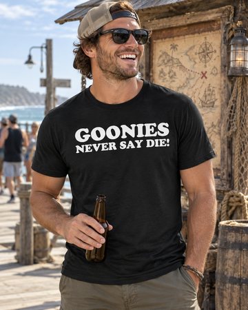 AI-generated lifestyle visualization: person wearing the black tri-blend Goonies Never Say Die! quote T-shirt in a weathered coastal boardwalk with an old treasure map kiosk and cool natural daylight
