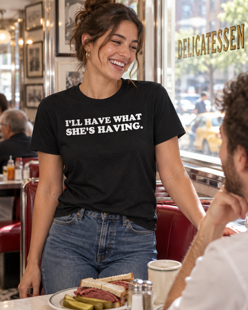 AI-generated lifestyle visualization: person wearing the black tri-blend I'll Have What She's Having. quote T-shirt in a classic New York delicatessen booth with chrome trim and bright midday window light