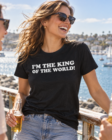 AI-generated lifestyle visualization: person wearing the black tri-blend I'm the King of the World! quote T-shirt in a sunlit harbor overlook with weathered railings and bright open sky