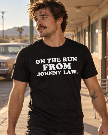 AI-generated lifestyle visualization: person wearing the black tri-blend On the Run from Johnny Law. quote T-shirt in a sun-bleached roadside motel walkway with centered doors, pale blue sky, and late-afternoon light