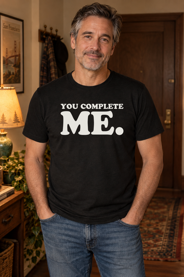AI-generated lifestyle visualization: person wearing the black tri-blend You Complete Me. quote T-shirt in cozy apartment entryway with warm evening light and understated 1990s decor