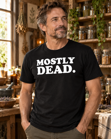 AI-generated lifestyle visualization: person wearing the black tri-blend Mostly Dead. quote T-shirt in cozy apothecary-style workshop with shelves of jars, rough wood, and warm window light