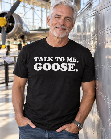 AI-generated lifestyle visualization: person wearing the black tri-blend Talk to Me, Goose. quote T-shirt in vintage aviation museum walkway with riveted metal walls and bright natural light