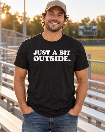 AI-generated lifestyle visualization: person wearing the black tri-blend Just a Bit Outside. quote T-shirt in a neighborhood baseball bleachers in late-afternoon sun with a scoreboard blurred in the distance