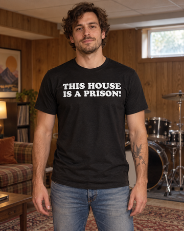 AI-generated lifestyle visualization: person wearing the black tri-blend This House Is a Prison! quote T-shirt in a suburban basement rec room with a drum kit in the background, wood paneling, and afternoon window light