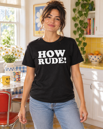 AI-generated lifestyle visualization: person wearing the black tri-blend How Rude! quote T-shirt in a bright 1990s kitchen breakfast nook with colorful mugs and soft window light