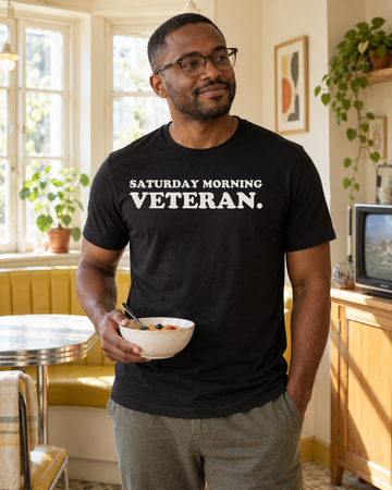 AI-generated lifestyle visualization of a person wearing the Saturday Morning Veteran. black tri-blend quote T-shirt in bright retro breakfast nook with a different adult man holding a cereal bowl near a generic small television, soft morning light, no visible brands, and the full blank black T-shirt front unobstructed