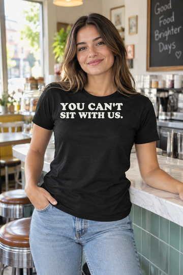 AI-generated lifestyle visualization of a person wearing the black tri-blend You Can't Sit With Us. quote T-shirt in bright neighborhood brunch counter with a different adult woman, candid seated pose, natural daylight, no franchise imagery, and the full black T-shirt front visible