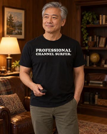 AI-generated lifestyle visualization of a person wearing the Professional Channel Surfer. black tri-blend quote T-shirt in relaxed wood-paneled den with a different adult man holding a generic remote, practical lamp light, no visible brands, and the full blank black T-shirt front unobstructed