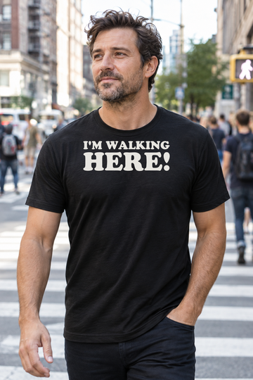 AI-generated lifestyle visualization of a person wearing the black tri-blend I'm Walking Here! quote T-shirt in busy urban crosswalk with a different adult man mid-stride, candid documentary framing, daytime light, no recognizable landmark, and the black T-shirt front clearly visible