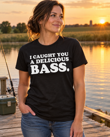 AI-generated lifestyle visualization: person wearing the black tri-blend I Caught You a Delicious Bass. quote T-shirt in quiet rural fishing dock at golden hour with a tackle box, still water, and open farmland