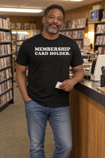AI-generated lifestyle visualization of a person wearing the black tri-blend Membership Card Holder. quote T-shirt in nostalgic video store aisle with a Black adult man holding a generic blank membership card, warm fluorescent light, no visible brands, and the full blank black T-shirt front unobstructed