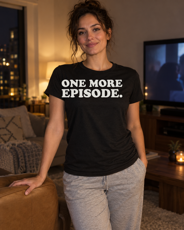 AI-generated lifestyle visualization of a person wearing the One More Episode. black tri-blend quote T-shirt in comfortable contemporary living room at night with a different adult woman curled in an armchair beside a dim generic television, cozy practical light, no visible brands, and the full blank black T-shirt front unobstructed