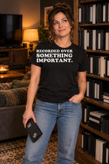 AI-generated lifestyle visualization of a person wearing the black tri-blend Recorded Over Something Important. quote T-shirt in comfortable home media room with an adult woman holding a generic blank tape, warm evening light, no visible brands, and the full blank black T-shirt front unobstructed