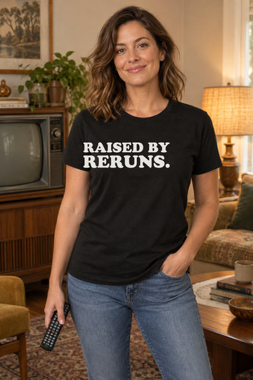AI-generated lifestyle visualization of a person wearing the black tri-blend Raised by Reruns. quote T-shirt in cozy living room with a different adult model holding a remote near a vintage television turned off, warm practical light, no logos, and the black T-shirt front unobstructed