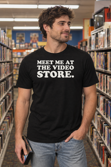 AI-generated lifestyle visualization of a person wearing the black tri-blend Meet Me at the Video Store. quote T-shirt in independent retro video rental aisle with a different adult woman browsing generic tape cases, fluorescent store lighting, no visible brands, and the black T-shirt front clearly visible