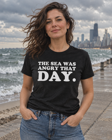AI-generated lifestyle visualization: person wearing the black tri-blend The Sea Was Angry That Day. quote T-shirt in windy city beach boardwalk under dramatic clouds with choppy water and muted 1990s color