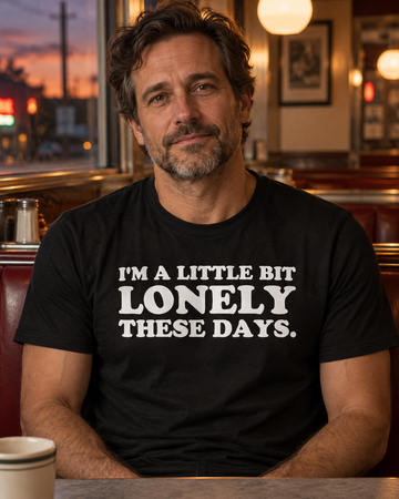 AI-generated lifestyle visualization: person wearing the black tri-blend I'm a Little Bit Lonely These Days. quote T-shirt in a quiet city diner booth at dusk with warm pendant lights and rain-softened windows