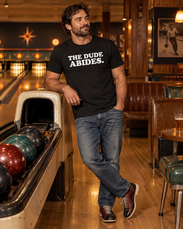 AI-generated lifestyle visualization: person wearing the black tri-blend The Dude Abides. quote T-shirt in a retro bowling alley lounge with amber lane lights and worn wood seating