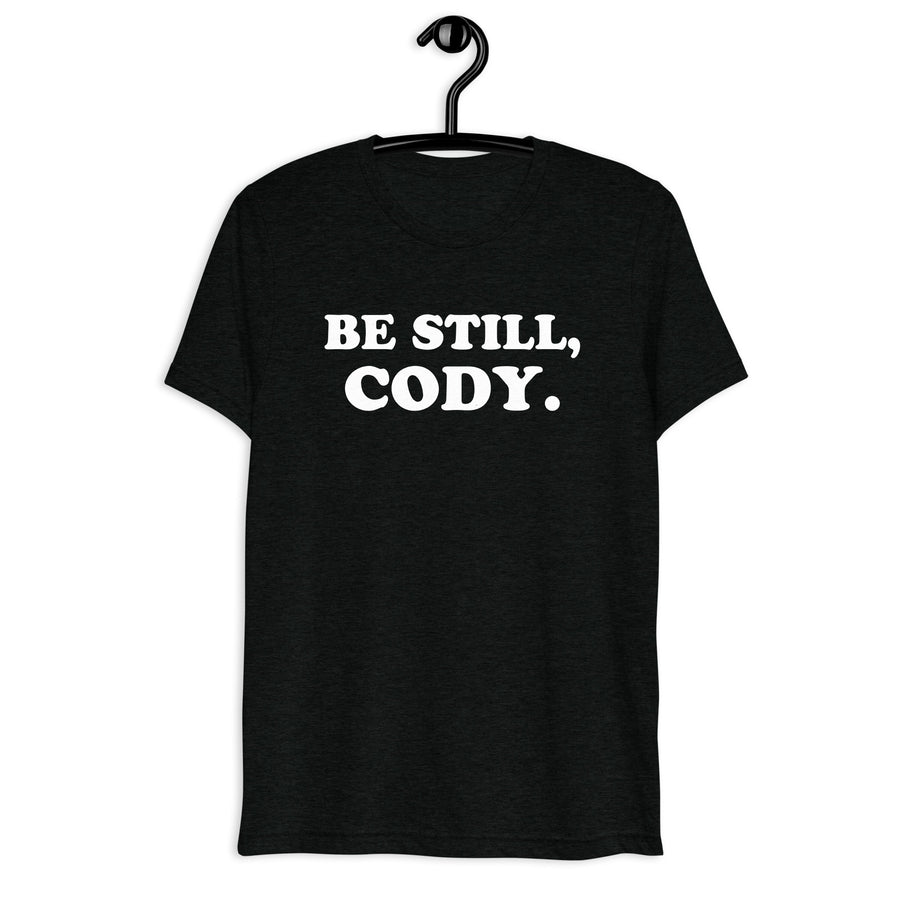 Front hanger view of black tri-blend quote shirt reading Be Still, Cody.
