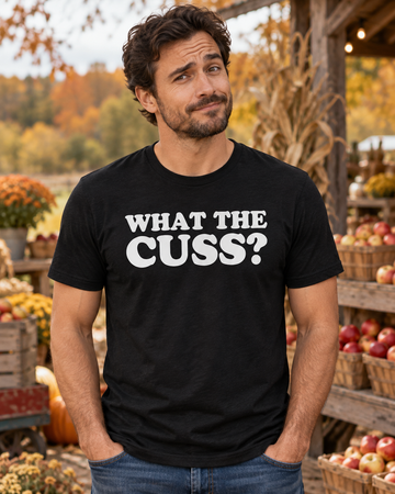 AI-generated lifestyle visualization: person wearing the black tri-blend What the Cuss? quote T-shirt in an autumn farm stand with apple crates, mustard-yellow siding, and crisp symmetrical composition